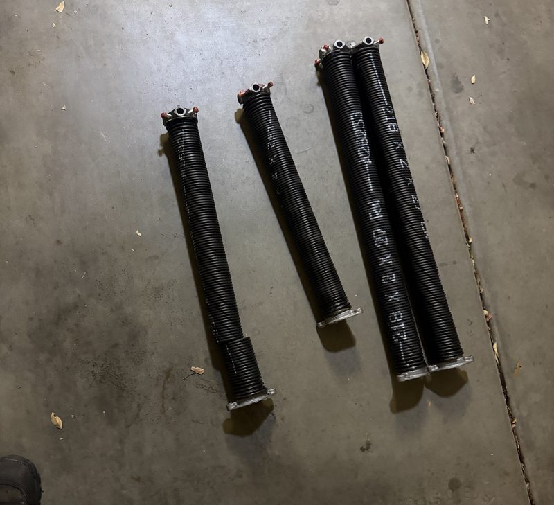 Four garage door torsion springs laid on a concrete floor, showing different lengths and sizes